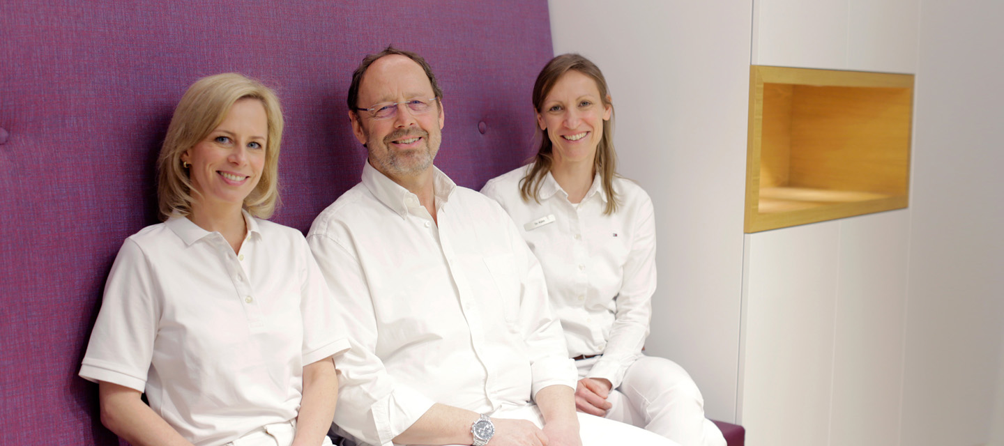 Dr. Schröder, Dr. Kühn und Dr. Böttcher sitzen nebeneinander im Flur und blicken freundlich in die Kamera.